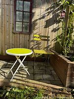 Round yellow top table with white metal legs beside weathered yellow painted chair in outdoor patio area showing outdoor wear.