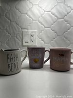 Three coffee mugs placed side by side on a white surface: 'PAPA BEAR' cream mug, 'BEST GRANDMA (EVER)' purple mug, and 'MAMA Bear' pink mug with bear image. Background is light-colored tile wall with outlet.