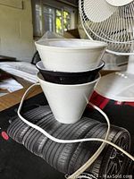 Three small conical lampshades stacked, two white and one black, placed on a black tire-patterned surface with a nearby white fan.