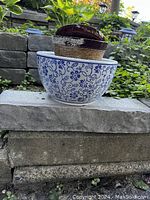 Photo of two plant pots placed on stone steps outdoors. One large white ceramic pot with blue floral vine designs and one smaller distressed painted pot with dark brown rim.