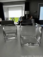 Photo showing two square glass vases on a white surface, one larger and upright, one smaller and tilted slightly.