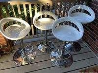 Set of 4 white plastic bar stools with chrome hydraulic lift bases and footrests, arranged on a porch deck.