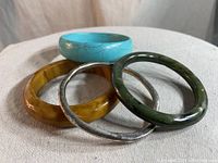Four vintage bracelets stacked on fabric surface. Includes turquoise, olive green, amber, and silver-tone bangles.