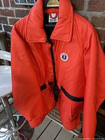 Front view of the bright orange Mustang survival jacket with a high collar, black adjustable waist strap, and zipper closure, showing overall condition and design.