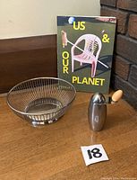 Alessi basket bowl, Michael Graves Lumina pepper mill grinder, and book Us and Our Planet shown together on wooden floor against wall.