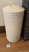 Cream colored tall ribbed Sally Designs trash bin with a domed lid, placed on wooden floor next to a brick corner