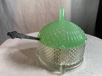 Side view of the sugar bowl showing clear textured crystal body with jadeite green glass lid and handle, and the metal spoon resting inside.
