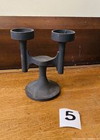Front view of a black cast iron 2-arm candelabra on a wooden surface with lot number 5 card visible.