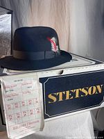 Black felt fedora style hat on original Stetson box, showing label with size 7 3/8, style 812, color black, with a black grosgrain ribbon band and multi-colored feather decoration.
