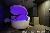 Flotation tank egg-shaped pod with lid open showing blue light inside in a tile floored room with chair and shower area.
