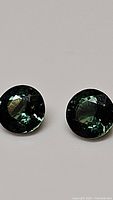 Close-up photo of two round cut sapphires with dark green and blue multicolored tones, clear facets and no visible inclusions.