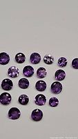 Multiple round cut, calibrated purple amethyst gemstones laid out on white background, showing various lighting reflections and the range of purple hues.
