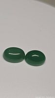 Photo of two oval green onyx cabochon gemstones placed side by side on a white background. The stones have a smooth polished, shiny surface.