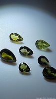 Seven pear cut green tourmaline gemstones arranged on white surface, with varying shades of green visible under focused light.