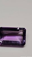 Top-down close-up of a rectangular emerald cut purple amethyst gemstone on a white background showing the color and cut details.