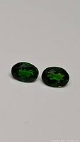 Front view of two oval cut, natural chrome diopside gemstones with electric green color resting on a white surface.