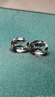 Two oval cut prasiolite gemstones lying on blue-green fabric, showing transparency and faceting.