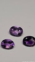 Three natural oval cut purple amethyst gemstones arranged on a white surface, showing color and cut details.