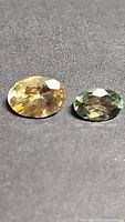 Close-up front view of two oval faceted tanzanite gemstones, yellow and green, placed side by side on a dark surface.