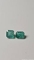 Photo displaying two rectangular emerald cut natural emerald gemstones on a plain background.