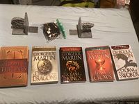 Five Game of Thrones books and two metal bookends with a sealed plastic bag of small collectibles arranged on a flat surface.