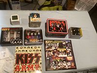 Overview of entire lot showing various boxed KISS collectibles including shot glasses, mini figures, tins, mug, and calendar.