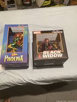 Two boxed Marvel Galleria series PVC diorama figures: X-Men Phoenix and Black Widow, front view on bed surface.