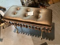 Front angle view of the bench showing the padded black tufted seat with tassel fringe on edges and white painted rod iron legs.