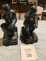 Two black ceramic statues of a seated man in thinking pose shown from front angle on a cloth surface.