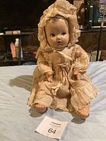 Full view of antique baby doll seated on a cloth surface, showing dress and bonnet details