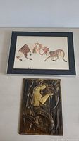 View of both artworks together on a flat surface: framed moose drawing and metal mermaid relief.
