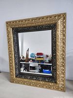 Front view of the antique gold gilt mirror showing the detailed leaf and scroll gilt outer frame surrounding a black ornate inner frame with carved floral motifs.