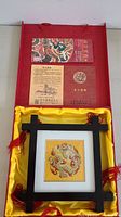 Top view of open red box with yellow satin lining, showing the framed cloth embroidery art piece with dragon and phoenix design and paperwork in lid.