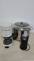 Lot arranged with white Braun coffee maker, large stainless steel cooking pot with closed lid, and black coffee grinder placed in front on a neutral surface.