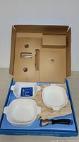 Overview of Corning Ware saucepan set in original blue box with cardboard inserts, two square white pans visible with use & care booklet.