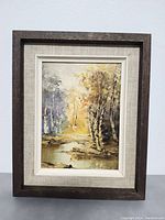 Framed oil painting showing a nature scene with birch trees and water, with visible textured brush strokes and earth tone colors.