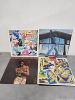 Photo of four vinyl records standing upright showing front cover artwork: General Public (colorful collage), Billy Joel (man jumping photo), Louie Louie Greatest Hits (shirtless man portrait), Level 42 (illustrated band members)