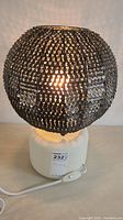 Vintage bronze-tone tassel table lamp illuminated, showing spherical shade texture and white base with attached power cord and inline switch.