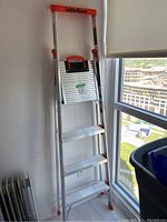 Photo of a folded aluminum ladder leaning against a wall near a window, showing the brand Little Giant on top with red accents and four steps visible.