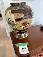 Front view of antique Japanese Satsuma vase showing floral and patterned decoration, sitting on wooden furniture with auction lot tag visible.