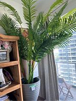 Artificial palm plant with green fronds in grey ribbed plastic pot next to wooden shelving and curtain.