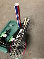 Photo of the badminton racket, cover, and a tube of birdies placed on the floor.