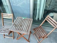 Photo showing the wooden bistro table and two chairs placed on a balcony with visible wear and weathering on wood surfaces.