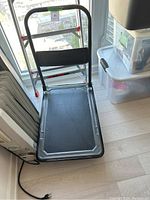 Folded view of the flatbed hand cart showing black anti-slip platform and foldable handle with metal frame and black rubber grip with red locking clips.