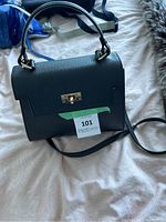 Front view of structured black genuine leather handbag with gold-tone twist lock, top handle and shoulder strap laid flat on a bed.