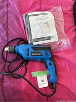 Blue and black Mastercraft electric hammer drill with attached power cord, laid on pink fabric next to instruction manuals in plastic bag.