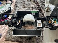 Top view of black compartmentalized nail kit box with open trays showing a white LED nail lamp and various nail accessories.