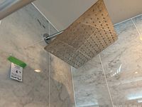 Square chromed rain shower head mounted on shower arm in bathroom with marble tile surround. Shower head face is 12x12 inches with evenly spaced nozzles.