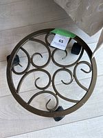 Top view of a round bronze metal plant stand with decorative scroll design and four black caster wheels.