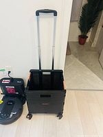 Open utility cart with black collapsible body and orange latches, with telescoping handle extended, positioned on light wood floor.